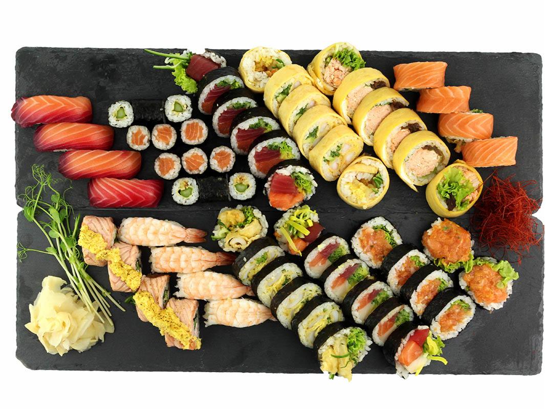 Elegancko ułożony zestaw sushi z różnymi rodzajami rolek, sashimi z łososia i krewetek na czarnej kamiennej desce, udekorowany imbirem i zielonymi pędami.
