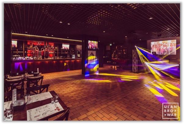 Wnętrze lokalu z barem, stolikami z menu i oświetleniem dyskotekowym, widoczny ekran z tańczącymi osobami.