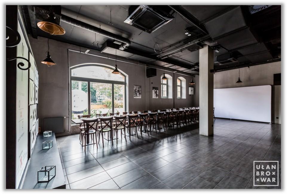 Sala bankietowa w stylu industrialnym z długim stołem nakrytym dla gości, widoczne okna z naturalnym światłem i dekoracyjne lampy wiszące