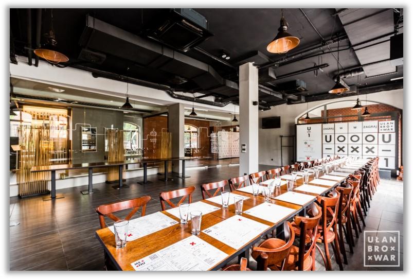 Długi, drewniany stół z nakryciami i szklankami w przestronnej sali restauracyjnej z czarnym sufitem i industrialnym oświetleniem.