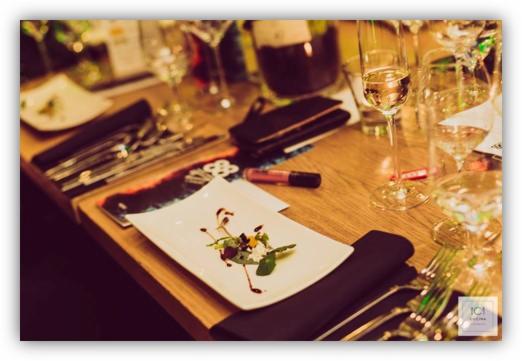 Elegancko zastawiony stół z kwadratowym talerzem z dekoracyjną kompozycją z ziół i sosem balsamicznym, obok kieliszek szampana, sztućce, portfel i butelka wina w tle.