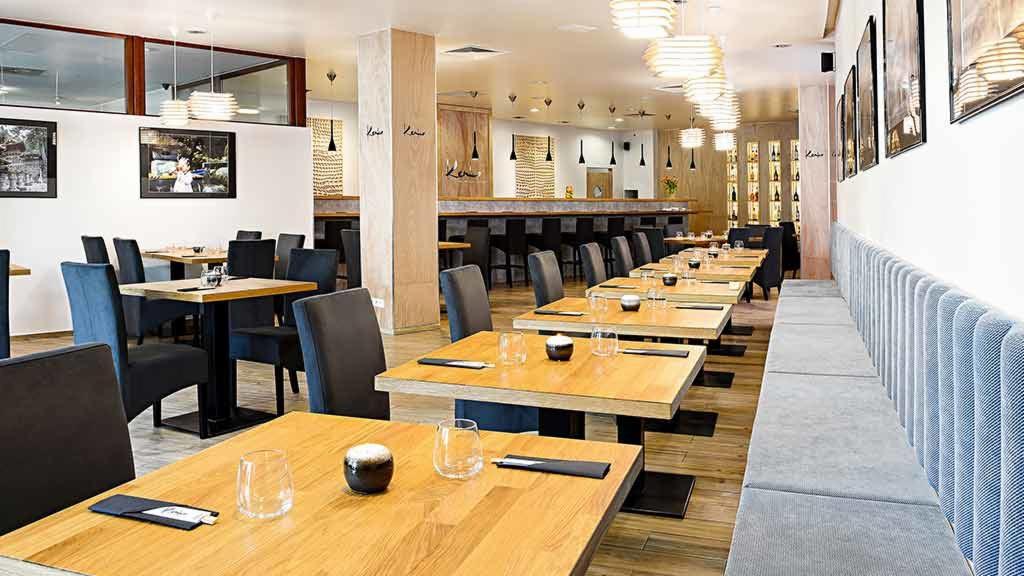 Jasne wnętrze restauracji z drewnianymi stołami nakrytymi szkłem i małymi czarkami, eleganckie krzesła i długa, pluszowa kanapa wzdłuż ściany, dekoracyjne lampy wiszące i artystyczne zdjęcia...