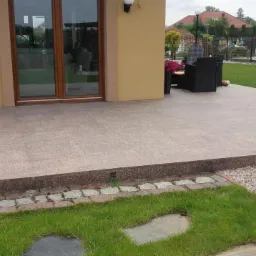 Wykończony taras z jasnych płyt, obrzeże z ciemnego granitu, widoczne meble ogrodowe i lampy oświetleniowe, trawnik z kostką i kamieniami ozdobnymi.