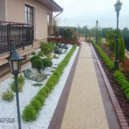 Ogród przydomowy z ścieżką z kostki, białymi kamieniami, lampami i równo przyciętymi krzewami wzdłuż ścieżki.