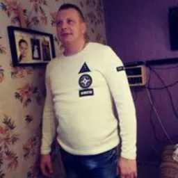 Mężczyzna w kremowym swetrze pozuje na tle ściany w kwiaty i fioletowej ściany z wiszącym sprzętem elektronicznym.