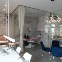 Nowoczesne wnętrze salonu urody z przeszkloną ścianą, widoczne stanowisko do manicure z lampą i frezarką, w tle fotel do zabiegów kosmetycznych i designerski żyrandol.