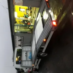 Pusta platforma lawety z rampą, zaparkowana przed warsztatem z żółtym oświetleniem wewnątrz, widok z boku na mokrym asfalcie w pochmurny dzień.