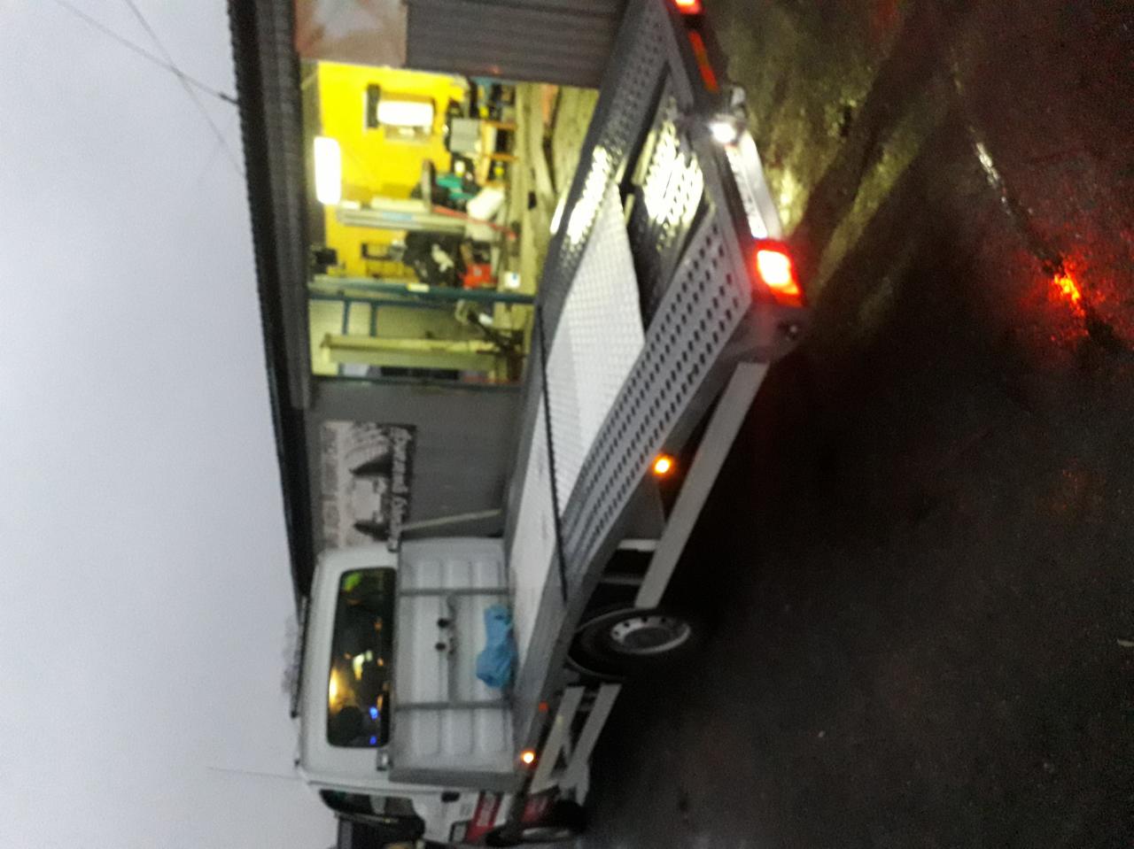 Pusta platforma lawety z rampą, zaparkowana przed warsztatem z żółtym oświetleniem wewnątrz, widok z boku na mokrym asfalcie w pochmurny dzień.