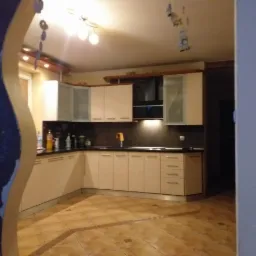 Widok na narożną, jasną zabudowę kuchenną z czarnym blatem, oświetleniem podszafkowym i ceramiczną podłogą, z widocznym fragmentem niebieskiej ściany z dekoracyjnym wykończeniem łuku.