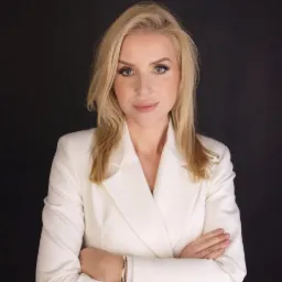 Portret kobiety w białym żakiecie na czarnym tle, z rękami skrzyżowanymi na piersiach i delikatnym uśmiechem.