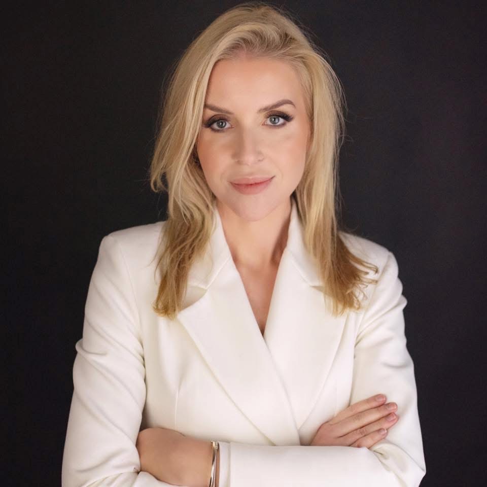 Portret kobiety w białym żakiecie na czarnym tle, z rękami skrzyżowanymi na piersiach i delikatnym uśmiechem.