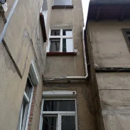 Zewnętrzna instalacja odprowadzająca wodę deszczową, wykonana z błyszczącej rury, zamontowana na elewacji budynku z widocznymi oknami i elementami instalacji elektrycznej.