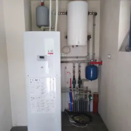 Węzeł cieplny w kotłowni: biały piec z wyświetlaczem i schematami, zbiornik akumulacyjny, naczynia przeponowe, liczne rury i zawory.