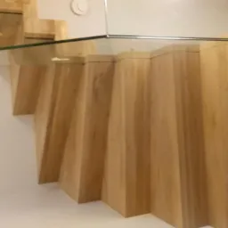 Nowoczesne schody z drewna z przezroczystym szklanym panelem, widok z dołu na stopnie o nietypowym, geometrycznym kształcie.