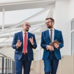 Dwóch mężczyzn w garniturach idzie korytarzem, jeden trzyma smartfon, drugi tablet, prowadząc rozmowę.