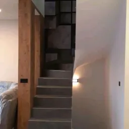 Nowoczesne schody z szarymi stopniami, drewnianą kolumną i dekoracyjną ścianą z kwadratowych elementów w odcieniach szarości, oświetlone kinkietem.