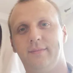 Portret mężczyzny w koszuli z wzorem, z lekkim uśmiechem i delikatnym cieniem na policzku.