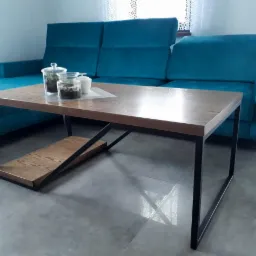 Stolik kawowy z drewnianym blatem i czarną, metalową podstawą, z dodatkową półką, na tle turkusowej sofy i szarych płytek podłogowych.