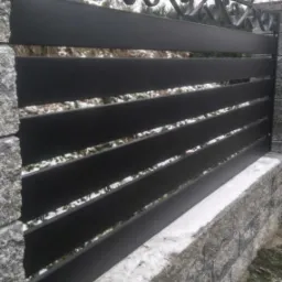 Nowoczesne, poziome przęsła ogrodzeniowe w kolorze czarnym, z dekoracyjnym, kutym elementem u g&oacute;ry, osadzone na kamiennym murku pokrytym śniegiem.