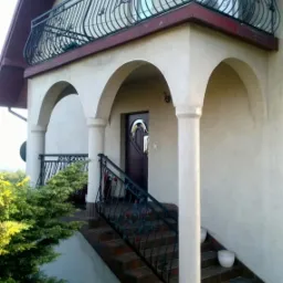 Dwukondygnacyjny dom z balkonem zabezpieczonym kutą, czarną balustradą z ozdobnymi elementami, widoczne schody wejściowe z poręczą w tym samym stylu, oraz roślinność w donicach.