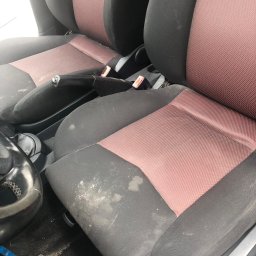 pranietapicerki-bydgoszcz - Brudne, czarno-czerwone fotele samochodowe z plamami i zabrudzeniami, widoczne elementy wnętrza auta, kierownica i dźwignia zmiany biegów.