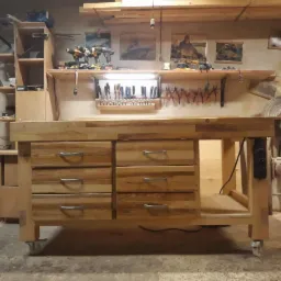 Solidny, drewniany stół warsztatowy z szufladami na kółkach, wyposażony w narzędzia i oświetlenie, stojący w warsztacie stolarskim.