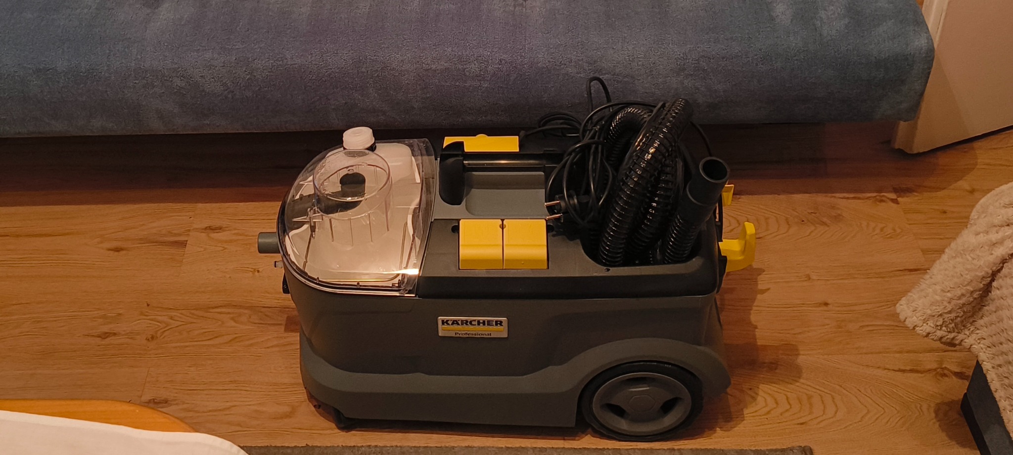 Profesjonalny odkurzacz piorący Kärcher na drewnianej podłodze, gotowy do użycia. Widoczny zbiornik na wodę i zwinięty wąż. W tle fragment sofy.