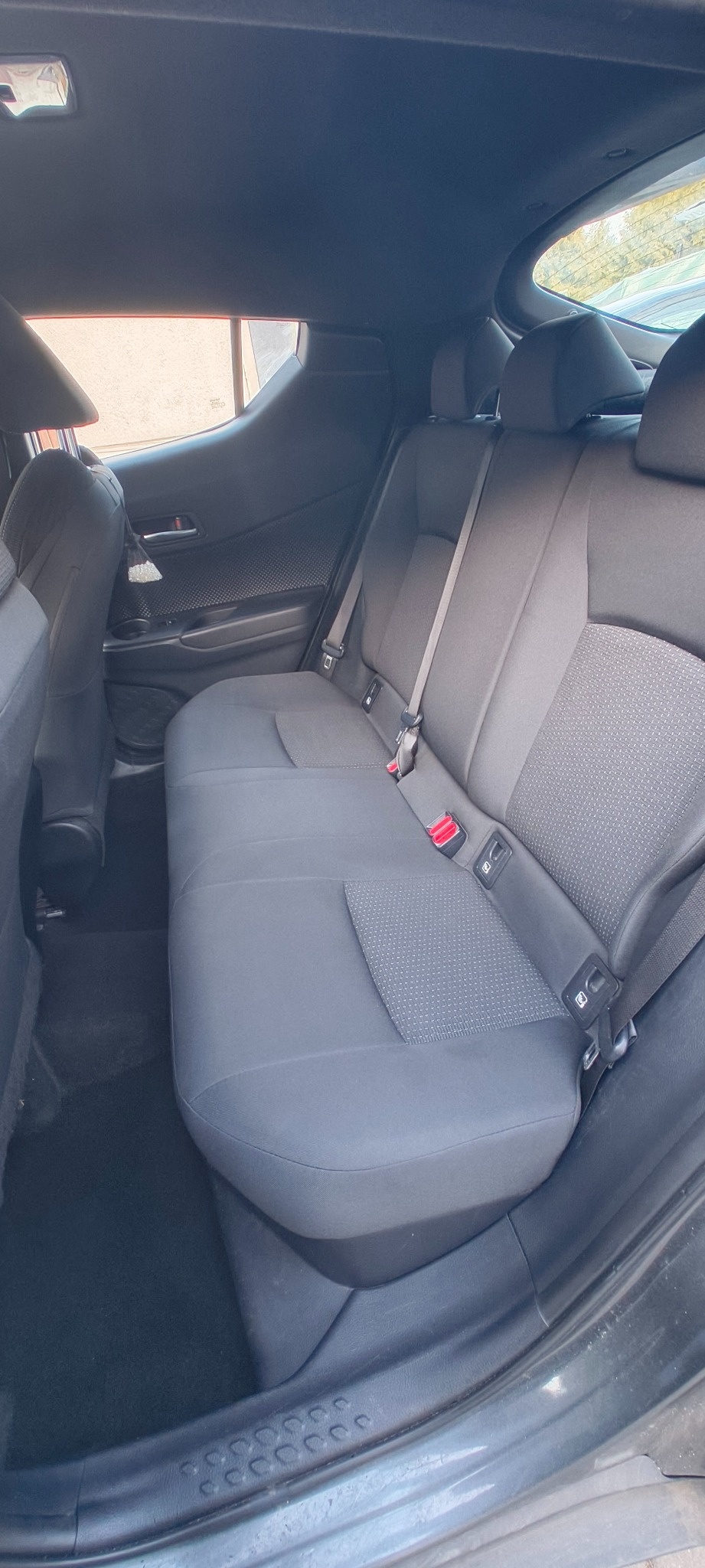 Wnętrze samochodu z widoczną tylną kanapą z szarej tapicerki. Widoczne pasy bezpieczeństwa i zagłówki. Ciemna podłoga i elementy wykończenia wnętrza.