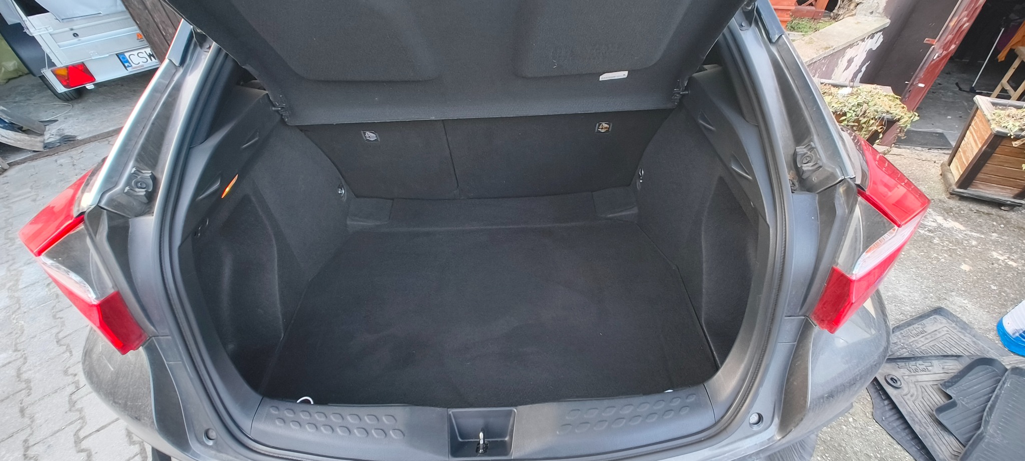 Otwarty bagażnik szarego samochodu typu hatchback z widoczną czarną tapicerką. W tle przyczepka samochodowa i elementy otoczenia.