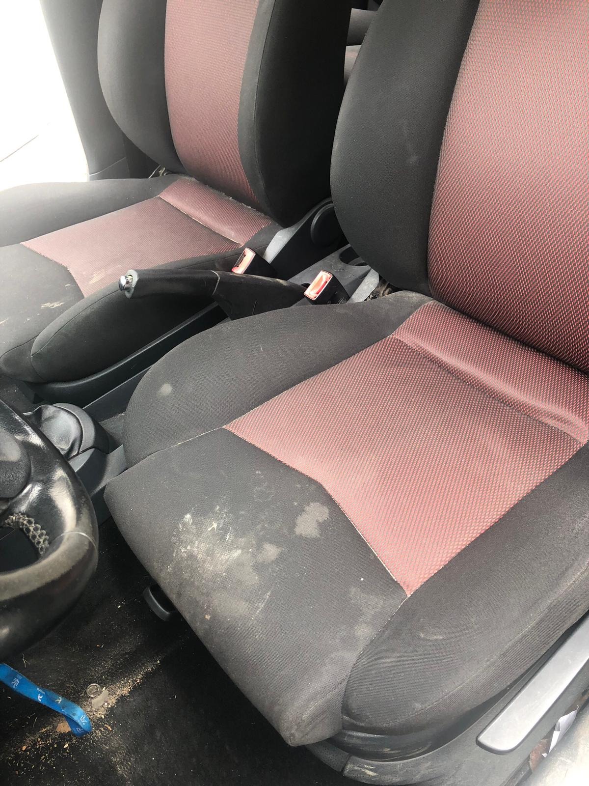 Brudne, czarno-czerwone fotele samochodowe z plamami i zabrudzeniami, widoczne elementy wnętrza auta, kierownica i dźwignia zmiany biegów.