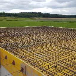 Zbrojenie stropu domu jednorodzinnego, widoczne żółte szalunki i stalowe pręty zbrojeniowe na tle zielonych pól i lasu.