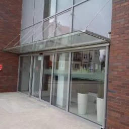 Szklane drzwi wejściowe do budynku z aluminiową ramą i szklanym zadaszeniem podtrzymywanym przez stalowe linki, widoczne rośliny w donicach za szybą i ceglana ściana z boku.