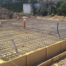 Siatka zbrojeniowa ułożona na żółtej izolacji w szalunku drewnianym, przygotowana do wylania fundamentów, z widocznymi wystającymi rurami instalacyjnymi.