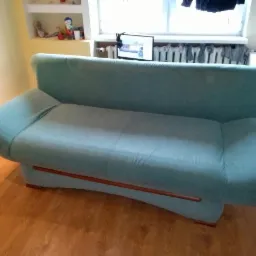 Turkusowa kanapa z drewnianymi elementami na tle pokoju z grzejnikiem i suszącymi się ubraniami.