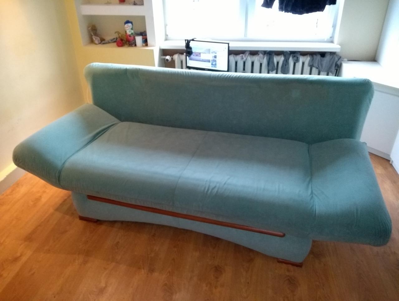 Turkusowa kanapa z drewnianymi elementami na tle pokoju z grzejnikiem i suszącymi się ubraniami.