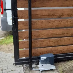 Automatyczna brama przesuwna z drewnianymi panelami, widoczny napęd NICE i lampa ostrzegawcza, świeżo po montażu, ziemia wokół napędu naruszona.