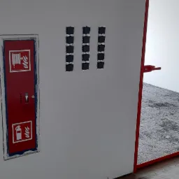 Biała ściana z trzema rzędami puszek elektrycznych podtynkowych, obok czerwona szafka hydrantowa i framuga drzwi.