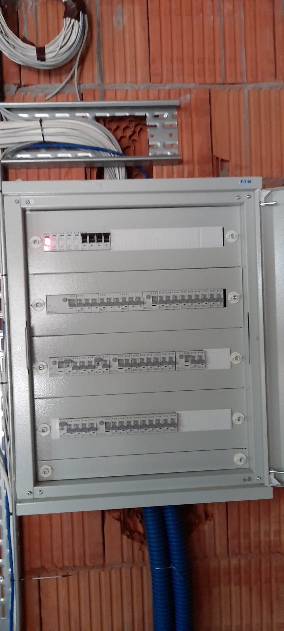 Otwarta skrzynka rozdzielcza firmy Eaton zamontowana na ceglanej ścianie, widoczne przewody elektryczne i niebieskie rury karbowane.