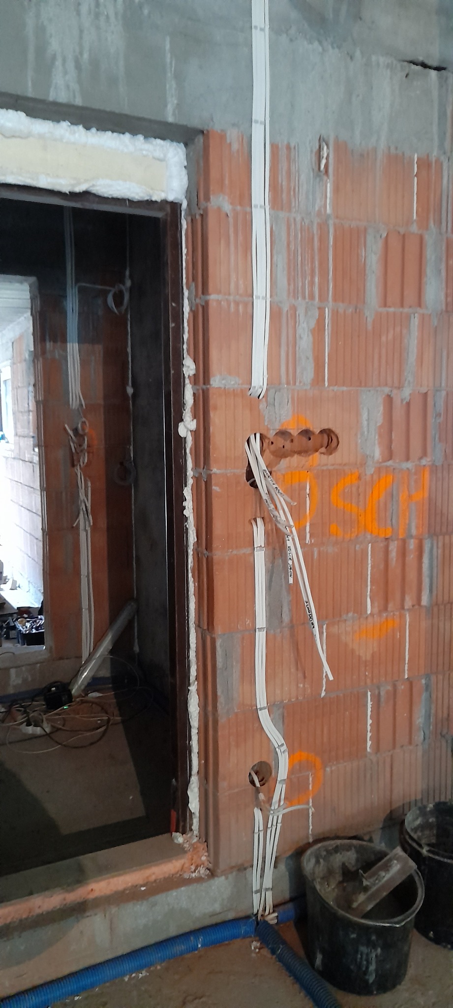 Surowa ściana z czerwonej cegły z wywierconymi otworami i poprowadzonymi kablami elektrycznymi w białych listwach, widoczne także fragmenty instalacji w otworze drzwiowym.
