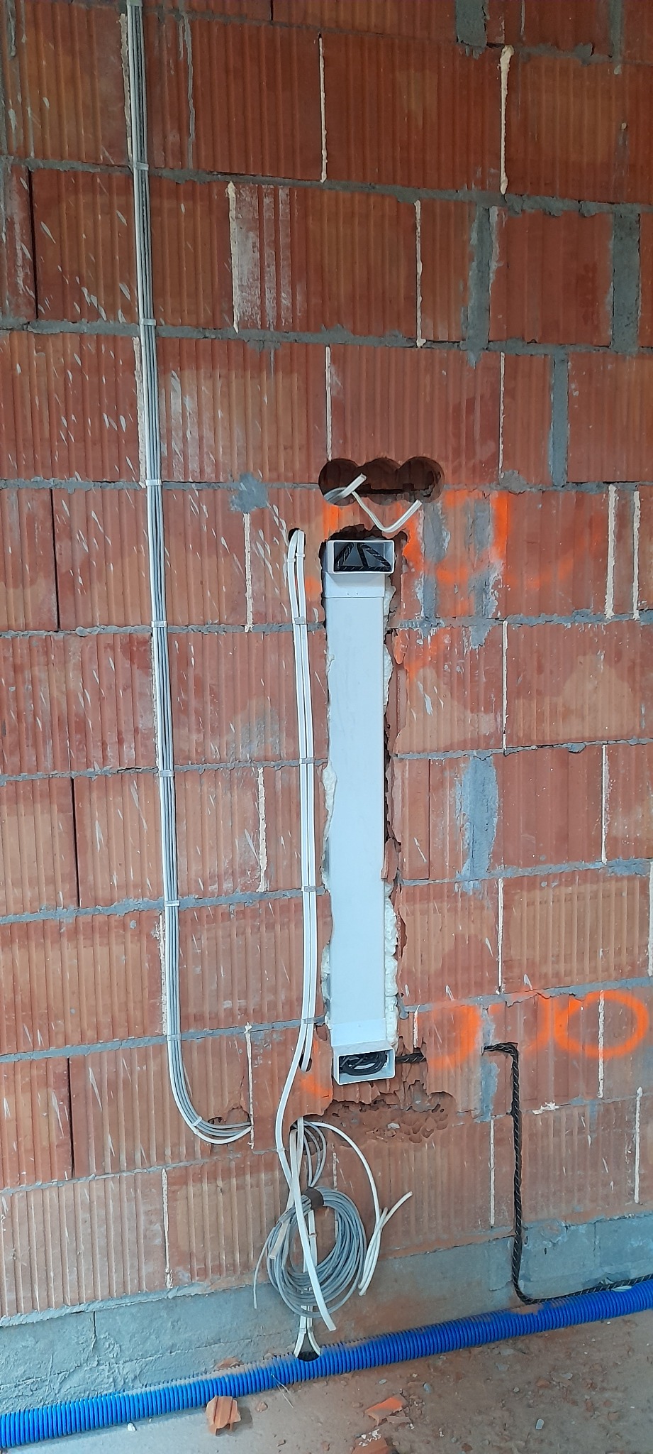 Ceglana ściana w trakcie prac elektrycznych: przewody w peszlu, puszka instalacyjna i kable zwisające z otworu w ścianie. Widoczne ślady po wierceniu i pomarańczowe oznaczenia na murze.