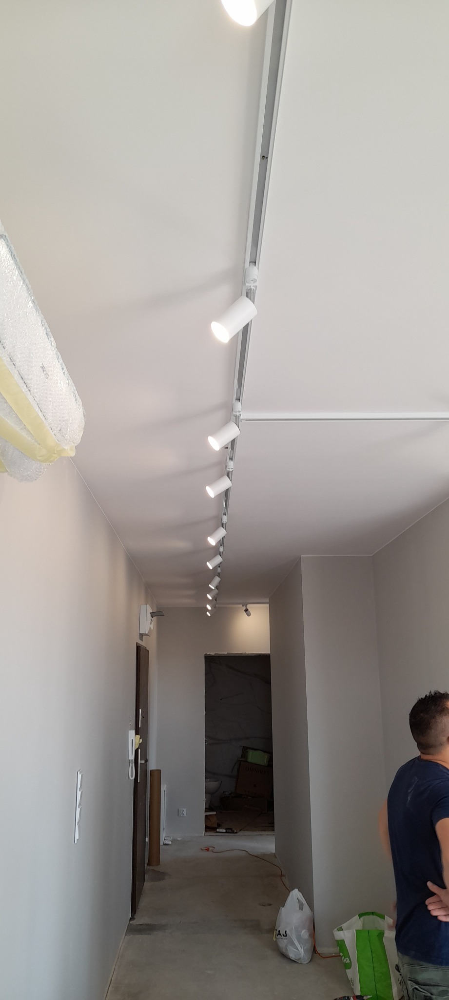 Zamontowane białe reflektory szynowe na suficie w korytarzu, widoczne ślady prac wykończeniowych.