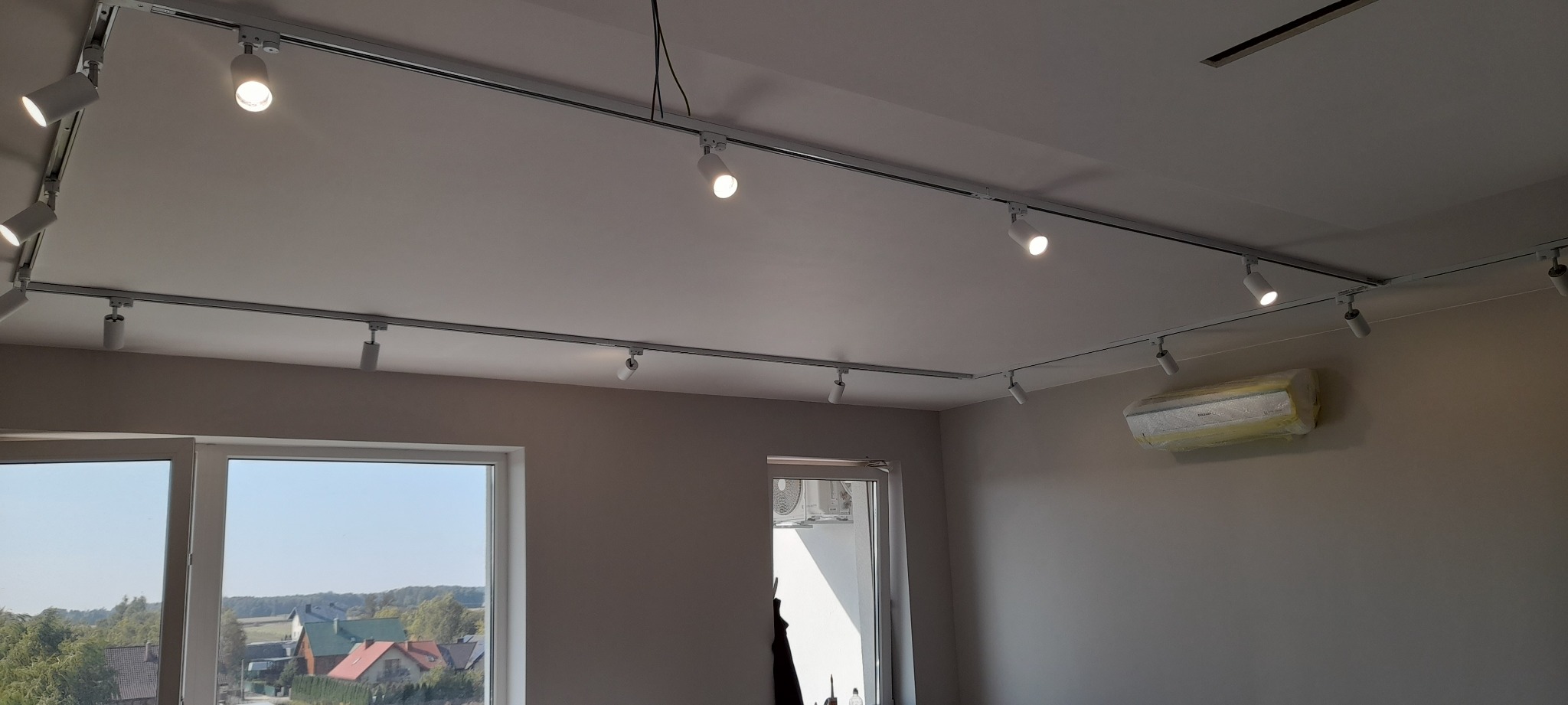 Białe reflektory szynowe zamontowane na suficie w jasnym pomieszczeniu, z widocznym oknem i klimatyzatorem zabezpieczonym folią.