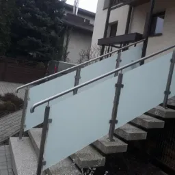 Zewnętrzne schody betonowe z balustradą ze stali nierdzewnej i szkła matowego, prowadzące do budynku mieszkalnego.