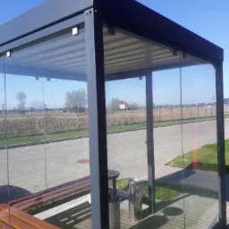 Zadaszona wiata przystankowa z metalową konstrukcją, szklanymi ścianami, drewnianymi ławkami i popielnicą, ustawiona przy chodniku i drodze w słoneczny dzień.