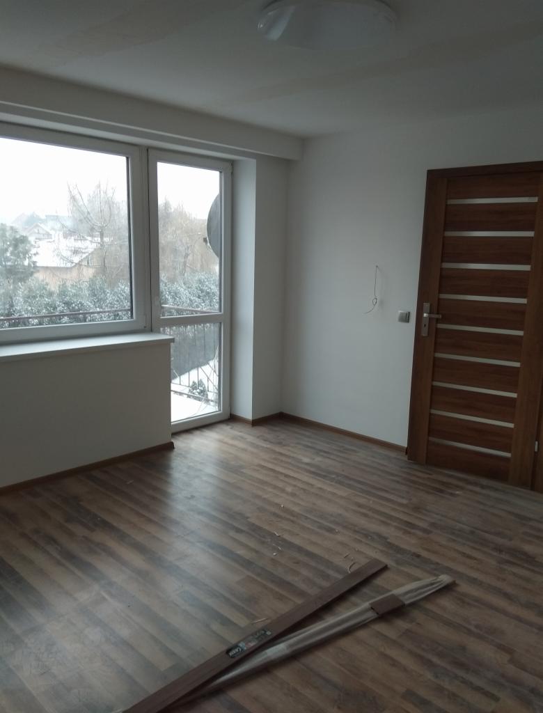 Pusty pokój z drewnianą podłogą w trakcie układania, widoczne listwy przypodłogowe w folii, okno z widokiem na zimowy krajobraz, drewniane drzwi wewnętrzne.