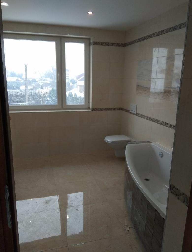 Jasna łazienka po remoncie z beżowymi płytkami, wanną obudowaną mozaiką i widokiem na zimowy krajobraz za oknem.