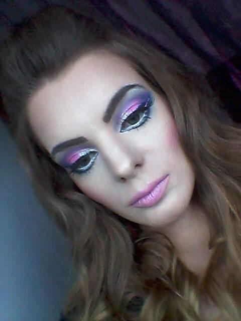 Portret kobiety z intensywnym, artystycznym makijażem oczu w odcieniach fioletu, różu i srebra, podkreślonym białym eyelinerem i sztucznymi rzęsami. Usta pomalowane na jasny róż, a policzki...