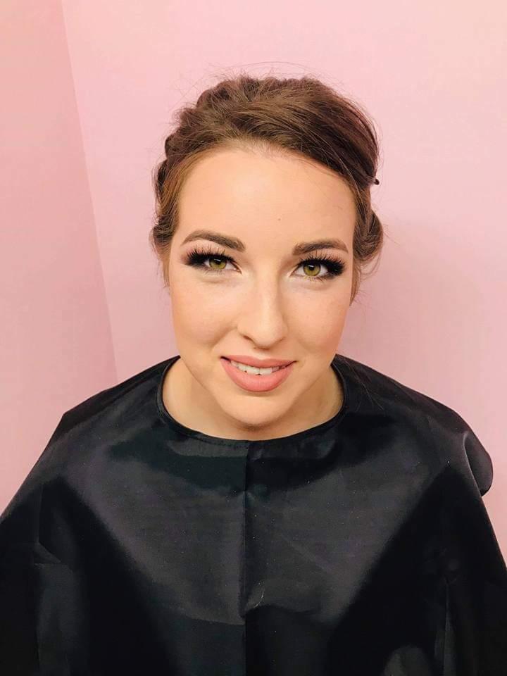 Portret kobiety z delikatnym makijażem, wyraźnie podkreślonymi rzęsami i upiętymi włosami, ubrana w czarny, satynowy fartuch kosmetyczny na tle pudrowo-różowej ściany.