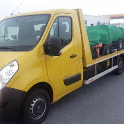 Żółta laweta Renault z przyczepą przewożącą sprzęt ogrodniczy, częściowo przykryty zieloną plandeką, na parkingu stacji benzynowej.