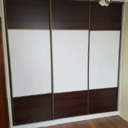 Trzydrzwiowa szafa przesuwna z ciemnobrązowymi i białymi panelami, widoczna lampa sufitowa z kloszami w kształcie kwiatów.